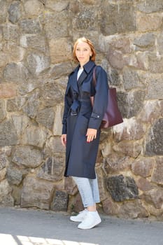 Full length portrait of redhead woman wearing trench coat, jeans and sneakers stands in front of stone wall