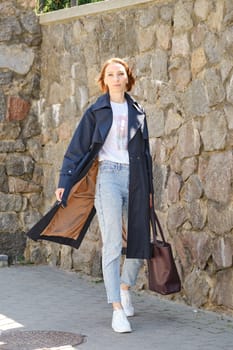 Elegant middle aged woman walks city street wearing blue trench coat, jeans and sneakers