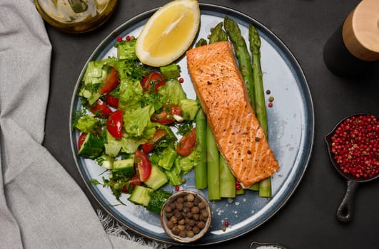 Healthy lunch with grilled salmon with asparagus and fresh tomato and cucumber salad, a row of lemon slice. Food on a blue round plate, top view
