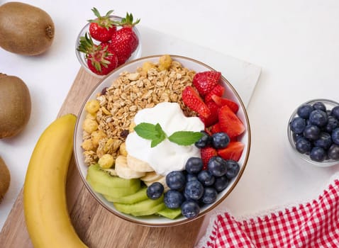 Granola with strawberries, kiwi, banana and blueberries in a round plate on a white table. Healthy and tasty food, top view