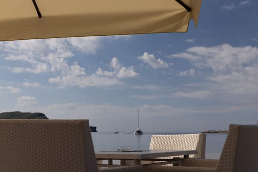 Sea view with table and chairs under a large umbrella. A sailboat can be seen in the distance under a cloudy sky. Copy space.travel or leisure content