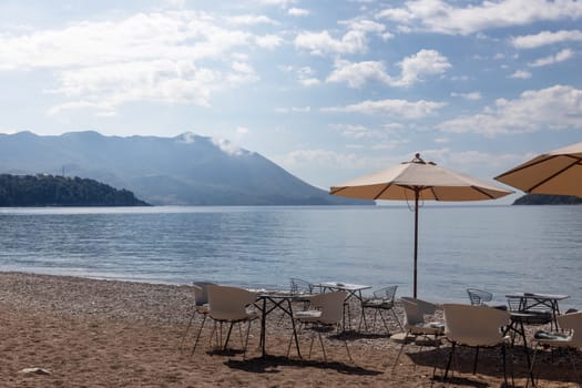 beach with tables and chairs under large umbrellas, with mountain background and cloudy sky. preparing for tourist season.travel or leisure content