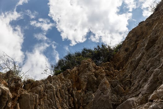 View from below of a rocky cliff with vegetation above against a blue sky and white clouds. Copy space.nature or abstract content