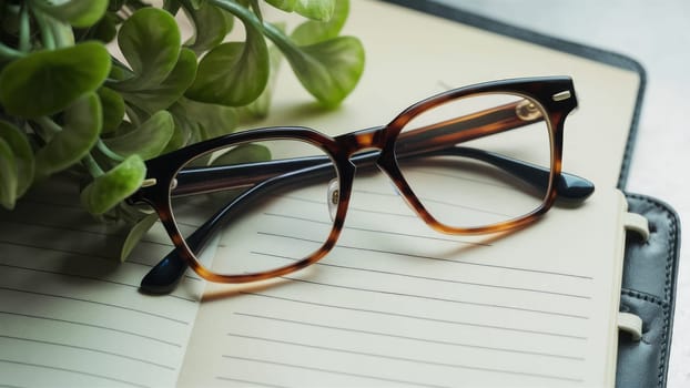 A pair of tortoiseshell glasses on an open notebook surrounded by a lush green plant, creating a calm and intellectual environment.