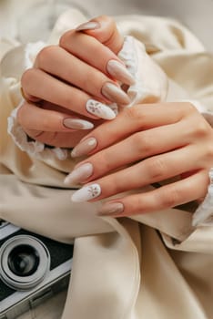 Close-up of female hands with elegant beige and white manicure with floral accents against a backdrop of silky fabric and a vintage camera.
