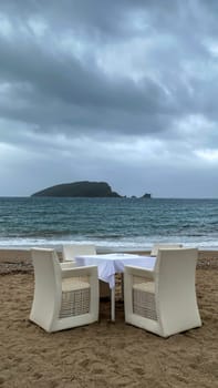 A table with a blue checkered tablecloth and white chairs set on a sandy beach, overlooking the turquoise sea and a distant island. Overcast sky adds a serene atmosphere. Copy space.