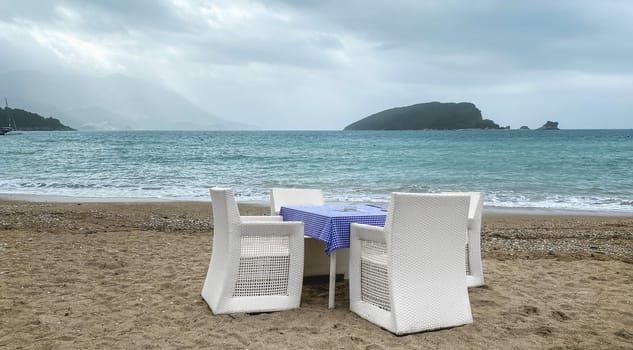 A set of white chairs and a table with a blue tablecloth on a sandy beach overlooking the sea and an island in the distance. The sky is overcast, adding to the moody atmosphere. Copy space.