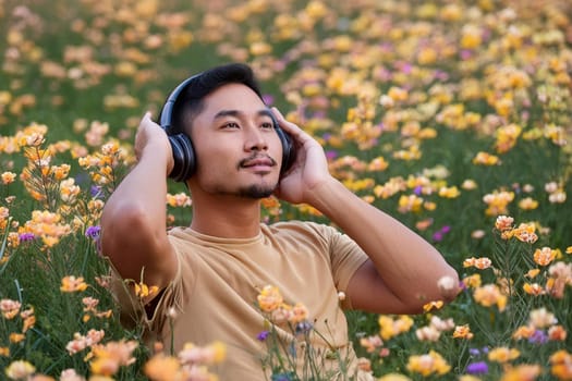 Asian man enjoying music in headphones sitting among wildflowers in meadow.