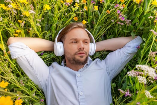 Man businessman in shirt and headphones lying on lawn among flowers and enjoying music during break or after work.