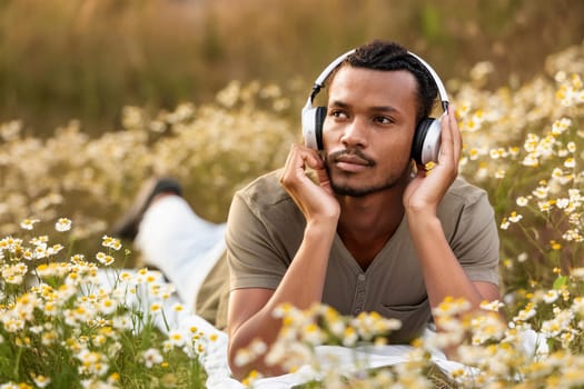 Black man lying in meadow among wildflowers and daisies and listening to music in headphones.