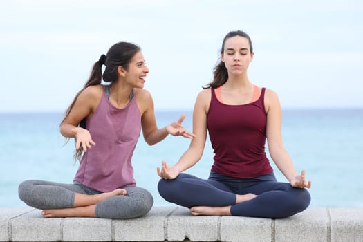 Front view portrait of a yogi trying to concentrate doing yoga and a friend disturbing on the beach