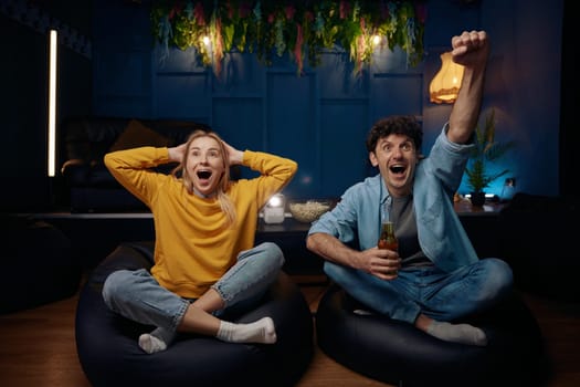 Impressed young couple watching TV in living room. Shocked man and woman rejoicing together