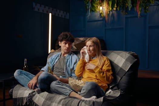 Man and crying woman watching emotional film with popcorn together. Young couple in living room spending time together