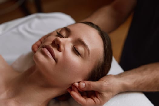 Closeup peaceful relaxed female face at massage session in spa center. Male masseur performing beauty procedure for facial skin, touching trigger points behind ears
