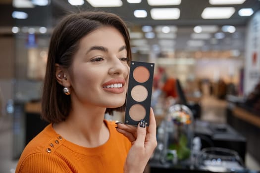 Woman customer choosing face blush holding it close to face. Lady in cosmetics shop looking for suitable make up products