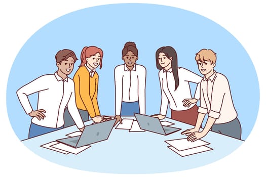 Team diverse business people stand at table with papers and laptops discussing company marketing strategy. Meeting in modern startup with team of young men and women engaged in corporate consulting