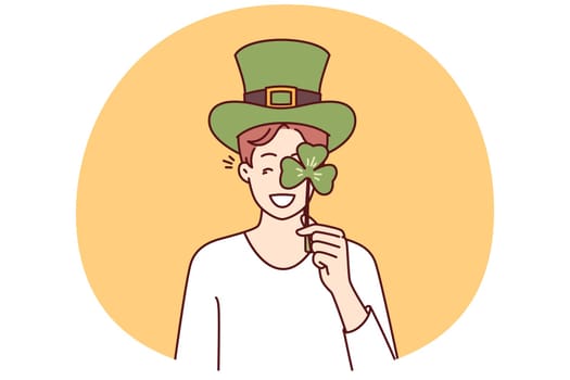 Happy man with St Patrick Day celebration accessories wearing green hat and covering eye with clover petal. Positive guy invites to traditional festival on eve of Irish holiday St Patrick Day