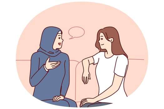 Girls from different ethnicities are talking sitting on couch demonstrating tolerance and lack of cultural prejudice. Woman in muslim clothes is talking to girlfriend of european appearance
