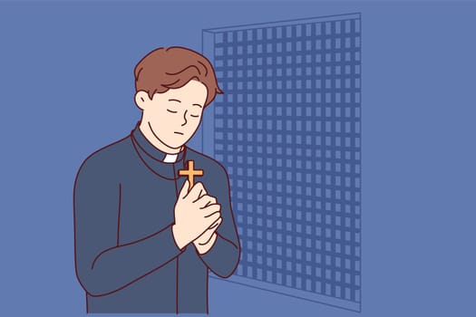 Padre prays and holds christian cross in hands, standing in confession room, asking god to atone for sins of temple visitors. Catholic priest observes ritual of confession and reads prayer