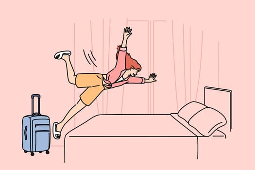 Happy woman tourist jumps on hotel bed, rejoicing at onset of vacation and opportunity to visit sunny resort. Girl with travel suitcase is in hotel and happily plops down on bunk to relax