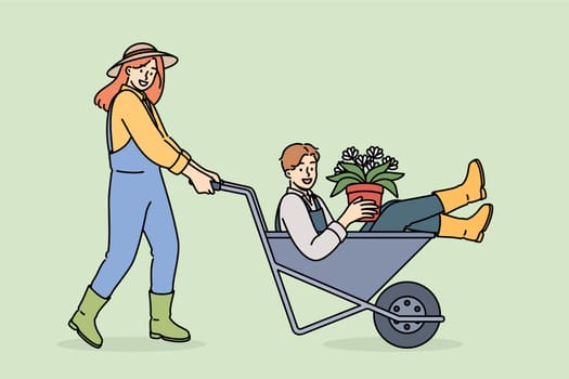 Woman farmer pushes man inside cart, enjoying good atmosphere in agro company team. Flowers in hands of farmer for planting seeds in fields and floriculture and getting large harvest