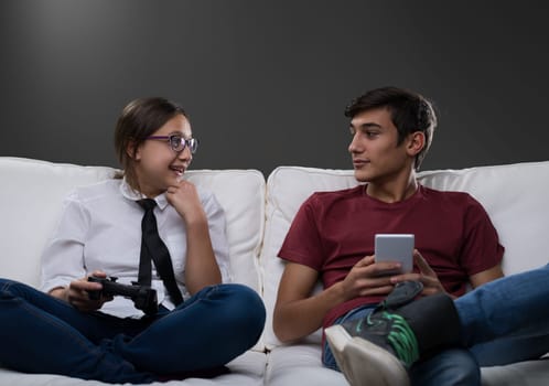 Two teenagers sit on a white couch discussing, their faces showing active engagement. The boy holds a smartphone while the girl, with a game controller, gestures, emphasizing collaborative education