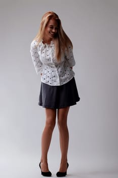 Confident and elegant young woman poses in a studio, smiling and looking down in a chic outfit. Her long blonde hair cascades down her back, exuding joy and positivity