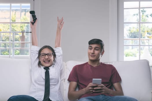 On a white couch, a girl celebrates her video game victory with arms raised, eyes closed in joy, as her brother watches with a composed and warm expression
