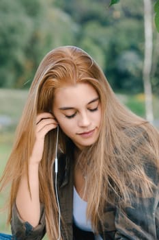 Young blonde woman listens to music outdoors, eyes closed, feeling peaceful and serene. She enjoys the tranquility of nature, lost in her playlist, blending technology and nature harmoniously
