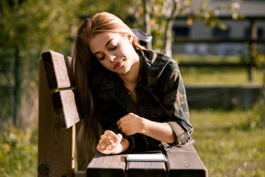 Young woman is lying on her stomach on a wooden bench outside, using a digital tablet. She is wearing a camouflage patterned shirt