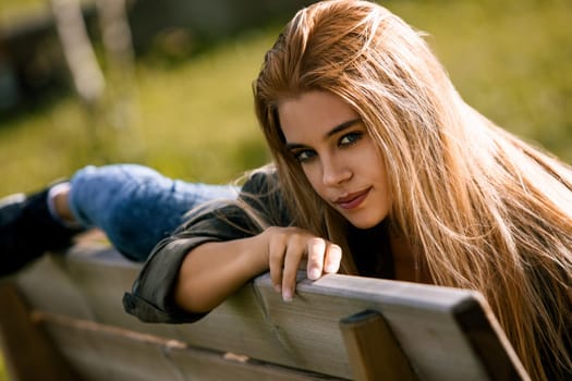 Young woman is lying on her stomach on a park bench, her long blonde hair cascading over her shoulders. She is enjoying the peace and quiet of nature
