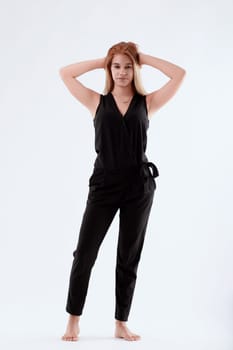 Stylish young woman in a black jumpsuit poses barefoot against a white background, exuding elegance and confidence as she holds her hair