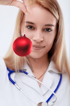 Joyful female doctor smiles holding a red christmas bauble, radiating happiness and festive cheer. Her blonde hair and white background highlight her professionalism and dedication to healthcare