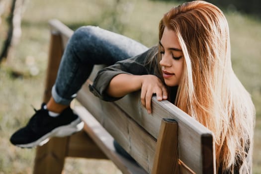 Young woman wearing casual clothing sits on a park bench, her legs stretched out in front of her. Her contemplative expression suggests a moment of quiet reflection