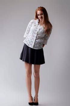 Confident young woman posing in stylish studio, exuding elegance with red hair cascading down her back, dressed in white blouse and black skirt, radiating fashion and style with cheerful smile