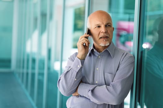 Mature businessman is speaking on his smartphone while leaning against a glass wall in an office building, demonstrating the empowering capabilities of modern technology
