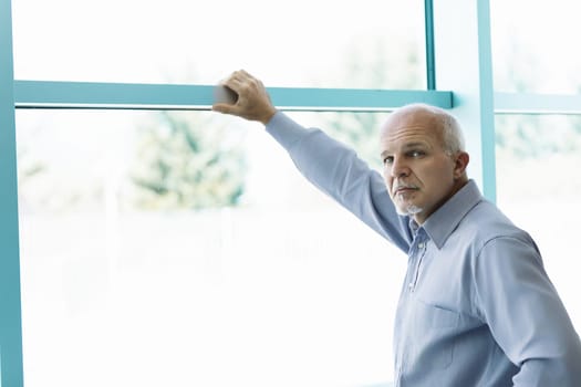 contemplative middle-aged man with a goatee, in a light blue shirt, leans on a large window frame. The outdoor background is brightly lit and softly blurred, enhancing his serious demeanor