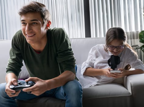 Two teenagers sitting on a couch, one intensely focused on playing a video game, holding a controller, while the other is engaged with her smartphone, both appearing content and relaxed