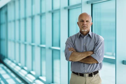 Confident senior businessman is posing with his arms crossed in a modern office building, representing experience and success in the business world