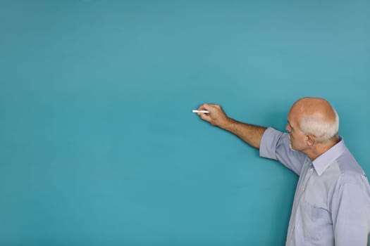 Bald man with a white goatee writes on a blue surface with chalk. Dressed in a light blue shirt, he is focused on his task, extending his arm towards the wall