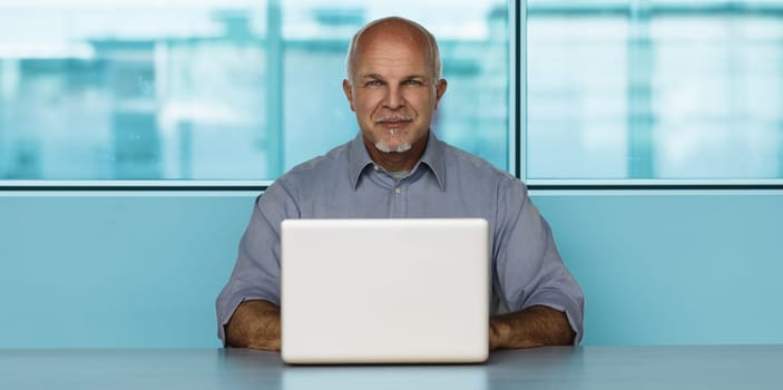 Mature businessman works on his laptop in a modern office space, exuding professionalism and success. Surrounded by a clean workplace with a city view, he appears efficient and productive
