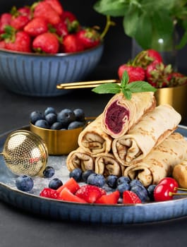 Crepes stacked in a pile, one of which is cut and filled with berry filling. The plate is also decorated with blueberries, strawberries, and cherries