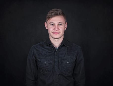 Portrait of a carefree young man against a black wall background looking to camera. Relaxed male model with joyful face expression wearing dark blue jeans jacket