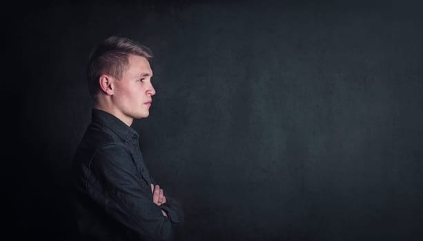 Pensive young man side view portrait, keeps arms folded against a dark wall background with copy space. Thoughtful male student with determined face expression