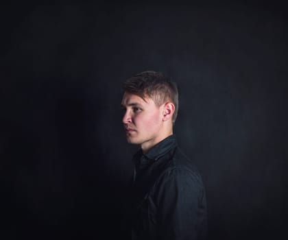 Thoughtful young man side view portrait, looks upset and tired against a dark wall background. Stressed male student feels emotional discomfort and anxiety problems