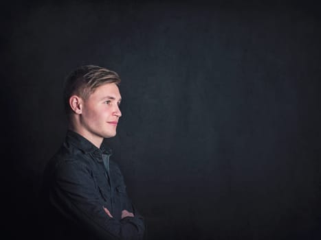 Positive thinking young man side view portrait, looks confident holding arms crossed against a black wall background. Male student reflecting with optimism