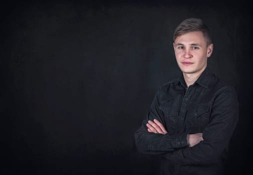Portrait of a confident young man, keeps arms folded against a black wall background with copy space. Successful male student with determined face expression wearing dark blue jeans jacket
