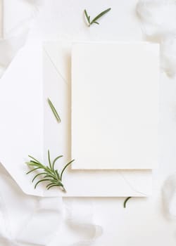 Blank cardand envelope near white silk ribbons and rosemary leaves top view, wedding mockup. Romantic scene with vertical cards and aromatic leaves flat lay. Valentines, Spring or Mothers day concept