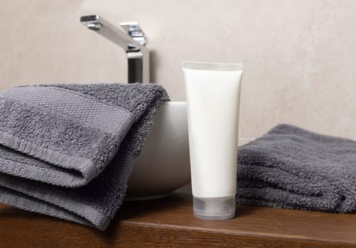 White cream tube near grey folded towel and vessel basin on brown wooden countertop in beige bathroom, close up, cosmetic mockup for brand packaging