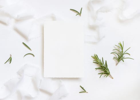 Blank card near white silk ribbons and rosemary leaves top view, wedding mockup. Romantic scene with vertical card and aromatic leaves flat lay. Valentines, Spring or Mothers day concept